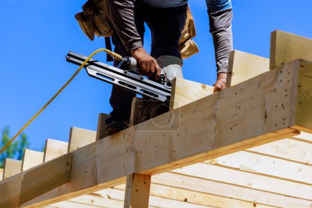 Téléchargez les photos : Travailleur de charpente installant des poutres à l'aide de clous d'air marteau dans le cadre en bois clouant - en image libre de droit