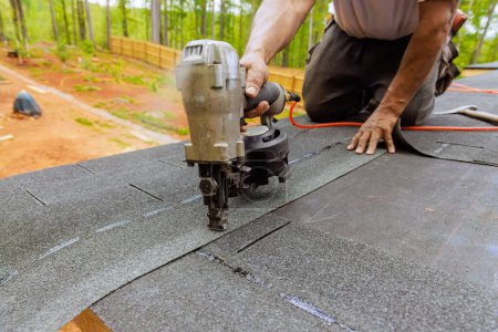 Using an air pneumatic nail gun, roofer install new asphalt bitumen shingles