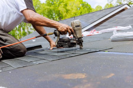 Photo for In addition to using an air pneumatic nail gun, roofer installs new asphalt bitumen shingles - Royalty Free Image