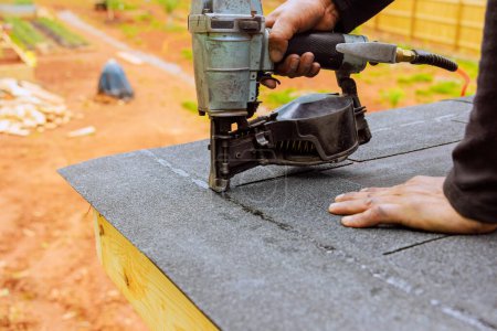 Roofer using an air pneumatic nail gun installs new asphalt bitumen shingles