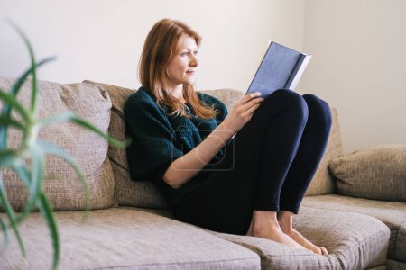 Téléchargez les photos : Beautiful mid aged Caucasian woman in casual clothes reading her diary relaxing in the living room. Hobby concept. - en image libre de droit