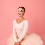 Adorable young woman in tutu with sincere smile on face posing tenderly over pink background. Skillful ballerina exercising alone in studio.