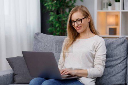 Attractive caucasian woman typing on wireless laptop while resting on couch. Mature lady wearing eyeglasses and casual clothes at home. Poster 620497976