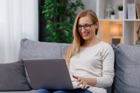 Happy caucasian woman in casual wear sitting on couch and looking on computer screen. Smiling mature blonde reading good news on wireless laptop. Poster 620498080