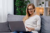 Happy caucasian woman in casual wear sitting on couch and looking on computer screen. Smiling mature blonde reading good news on wireless laptop. Poster #620498080