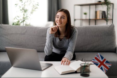 Attractive female using laptop and textbook for education while sitting on sofa at home. Beautiful young woman smiling happily and thoughtfully looking aside.