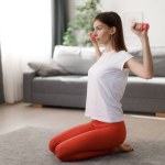 Attractive sporty woman in activewear sitting on floor and using dumbbells for training arms at home. Concept of domestic workout and healthy lifestyles.