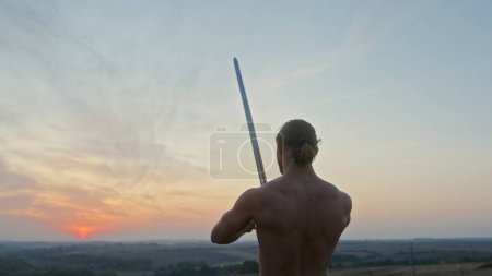 Hombre musculoso sin camisa con espada que practica movimientos de corte en una vista de ángulo bajo contra el cielo del atardecer en un lado en la vista trasera de ángulo bajo.