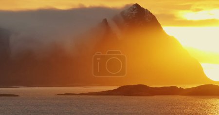 Sartén en tiempo real derecha calma que refleja el agua de mar cerca de la montaña borrosa y árboles verdes en colores anaranjados atardecer en las islas Lofoten Noruega Europa