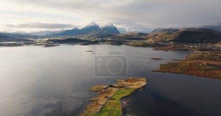 Real time zoom in aerial view of shining seawater with floating snow plates near green farming fields and houses on mountain ridges in Lofoten islands Norway under sky in daylight