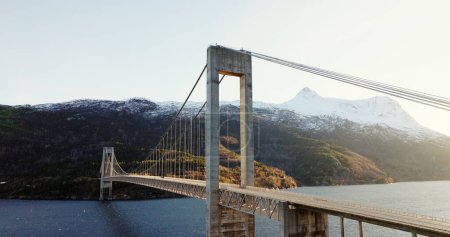 A suspension bridge gracefully stretches across a serene body of water, surrounded by mountains under the soft glow of the setting sun.