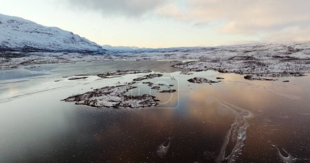 Real time zoom in aerial view of frozen seawater with floating ice flakes and snow capped mountain in Lofoten islands Norway under cloudy blue sky in daylight