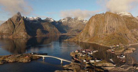 Real time zoom out aerial view of shining rippling seawater with way bridge and boats while houses near rocky snow capped mountains in Lofoten islands Norway under cloudy blue sky
