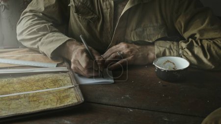 Soviet Army soldier analyzes map and draws up action plan sitting in abandoned house at table near window