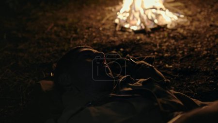 Soldier sleeping on the ground near a blazing campfire at night in a close up view as he turns restlessly under his blanket.