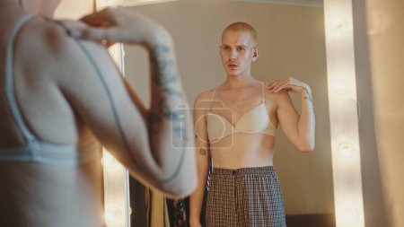 Real time handheld shot of young crossdresser putting on beige bra while dressing up in front of mirror with lamps