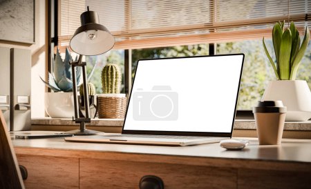 Téléchargez les photos : Un ordinateur portable avec un écran blanc se trouve sur un élégant bureau en bois dans un intérieur de style loft, avec des espaces verts en arrière-plan visibles par la fenêtre - en image libre de droit