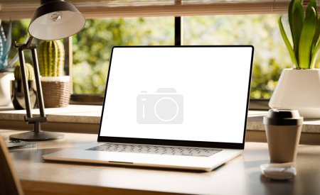 Téléchargez les photos : Un ordinateur portable avec un écran blanc se trouve sur un élégant bureau en bois dans un intérieur de style loft, avec des espaces verts en arrière-plan visibles par la fenêtre. 3d rendu - en image libre de droit