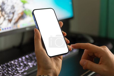 Téléchargez les photos : Un homme utilise un smartphone avec un écran vierge et sans cadre sur le fond d'un moniteur d'ordinateur. - en image libre de droit
