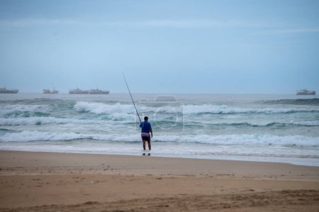 Téléchargez les photos : Man seen fishing with trawlers in distance - en image libre de droit