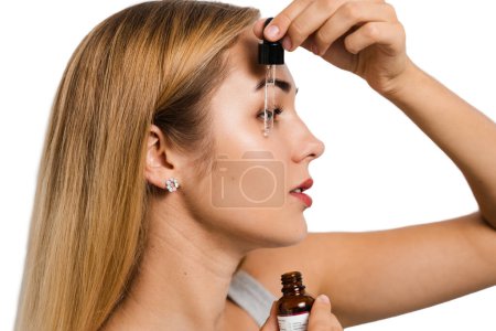 Téléchargez les photos : Jeune femme avec pipette sérum visage pour le traitement de l'acné et lissage des rides. Fille applique sérum visage sur son visage avec pipette sur fond blanc - en image libre de droit