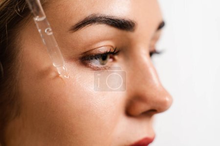 Photo for Girl drips facial serum on her cheek for acne treatment and wrinkle smoothing. Girl applies face serum to her face with pipette on white background - Royalty Free Image