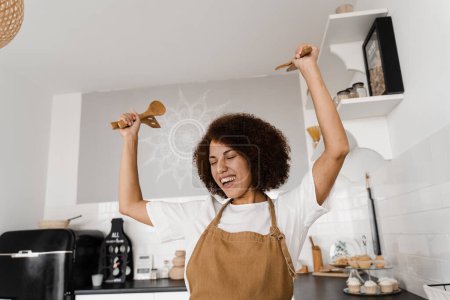 Foto de Mujer afroamericana está bailando en la cocina y levantando las manos. Chica africana cocinera en delantal bailando en la cocina blanca. Publicidad para cursos de cocina y talleres de comida - Imagen libre de derechos
