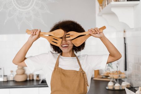 Foto de Ama de casa africana en delantal está posando con espátulas para cocinar en la cocina. Chica afroamericana cocinera divirtiéndose en la cocina. Publicidad para cursos de cocina y talleres de comida - Imagen libre de derechos