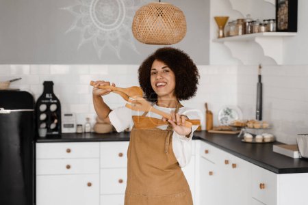 Foto de Chica africana cocinera en delantal bailando en la cocina blanca. Publicidad para cursos de cocina y talleres de comida. Mujer afroamericana está bailando en la cocina y levantando las manos - Imagen libre de derechos