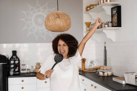 Foto de Ama de casa afroamericana en delantal cantando con espátulas para cocinar micrófono en la cocina. Joven africana alegre divirtiéndose con utensilios de cocina - Imagen libre de derechos
