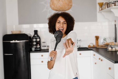Foto de Jovencita africana alegre divirtiéndose con utensilios de cocina. Ama de casa afroamericana en delantal cantando con espátulas para cocinar micrófono en la cocina - Imagen libre de derechos