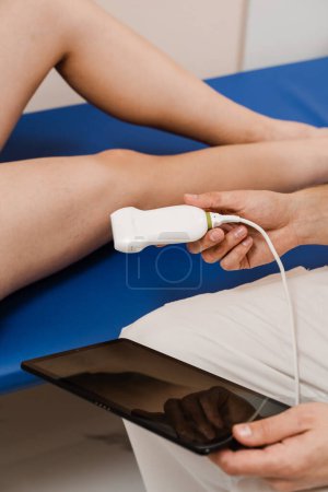 Photo for Vascular surgeon examines veins and arteries of legs using ultrasound machine of woman in medical clinic. Portable ultrasound machine for ultrasound legs scanning - Royalty Free Image
