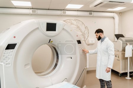 Foto de El médico del hombre ajusta el escáner de tomografía computarizada por TC en la clínica médica. Radiólogo barbudo guapo en bata médica con tomógrafo computarizado para obtener imágenes internas detalladas del cuerpo del paciente - Imagen libre de derechos