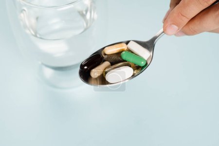 Téléchargez les photos : Pills on spoon of dietary biologically active supplements and glass of water for daily tablets intake on blue background. Set of omega and zinc pills, antioxidants from aging, lecithin and adaptogen - en image libre de droit