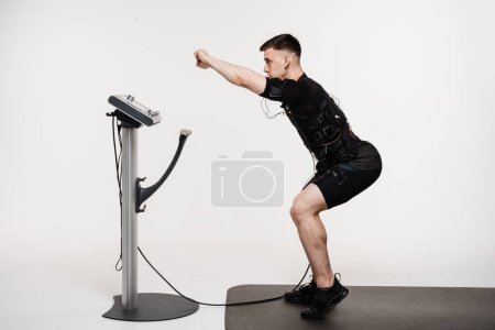 Photo for Muscular male athlete does squats physical exercises in EMS suit that uses electrical impulses to stimulate muscles on white background. Sport training in electrical muscle stimulation suit - Royalty Free Image