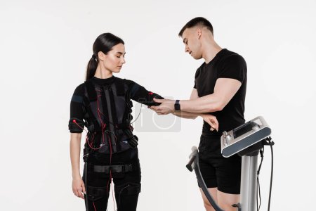 Photo for Trainer puts on girl EMS suit and checks contactors before training on white background. Sport group training in EMS electrical impulse muscle stimulation suit - Royalty Free Image
