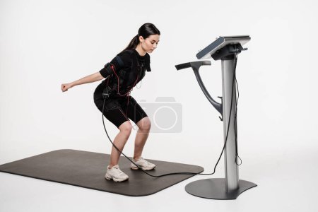 Photo for Sport training in EMS electrical muscle stimulation suit. Fitness girl in EMS suit is doing workout sport training that uses electrical impulses to stimulate muscles on white background - Royalty Free Image