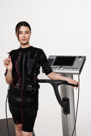 Photo for Fit girl in EMS suit with contactor in hand that uses electrical impulses to stimulate muscles on white background. Sport training in electrical muscle stimulation suit - Royalty Free Image