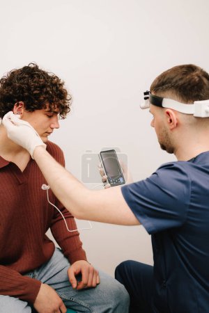 Téléchargez les photos : Le médecin ORL procède au contrôle de l'oreille du patient masculin avec un otoscope numérique. Homme patient et médecin ORL regardent l'écran d'un otoscope numérique lors d'une consultation - en image libre de droit