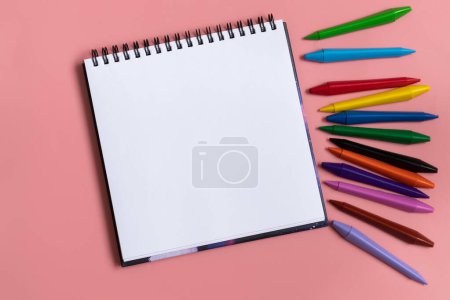 Foto de Cuaderno abierto con blanco vacío en blanco. Lápices de cera de colores sobre el fondo rosa pastel. Composición laica plana - Imagen libre de derechos