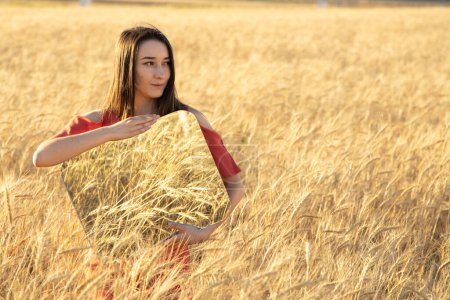 Photo for Smiling Young woman in the wheat field, holding mirror glass where reflected the dry wheat. Meditation, mental health concept. Copy space. - Royalty Free Image