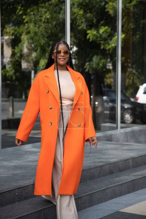 Foto de Una mujer afroamericana de moda en un abrigo de cachemira naranja y gafas de sol en una calle de la ciudad - Imagen libre de derechos