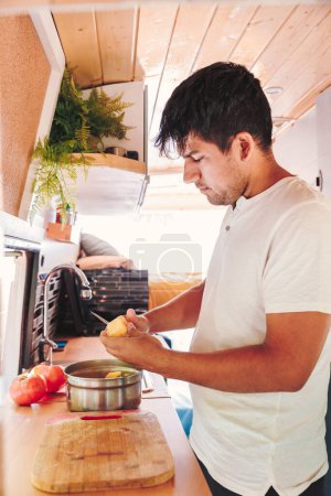 Young man preparing meal in the kitchen of his camper van. Van road trip holiday and outdoor summer adventure. Nomad lifestyle concept