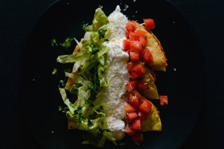 Photo for Mashed potato and chorizo fried tacos dorados with lettuce, tomatoes, sour cream and cotija cheese. Shot on black background. - Royalty Free Image