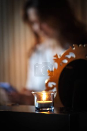 Una llama parpadeante de una vela en un vaso ilumina la mesa, emitiendo tintes y sombras. En el fondo, una mujer es apenas visible en la oscuridad de la habitación durante un evento
