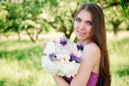 Photo for Pretty gentle young girl in a purple dress in garden with a bouquet in her hands looking at the camera. Professional makeup and dress for bridesmaids in violet. Copy space. - Royalty Free Image