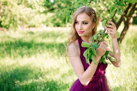 Photo for Professional model with makeup and hairstyle poses in a green garden and holds an apple tree branch in a fashionable dress. Beauty shot and copy space. - Royalty Free Image