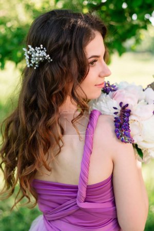 Photo for Beautiful girl in a purple dress stands with her back to the camera and poses with a bouquet of peonies in her hands. Curly hair with hairstyle and decoration. High quality shot. - Royalty Free Image