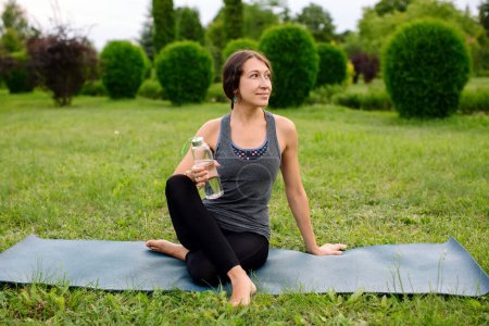 Photo for Young woman in comfortable sportwear sitting left twist on yoga mat with a water bottle relaxed after workout outside in the green park. High quality shot. - Royalty Free Image