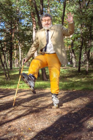 Foto de El viejo gracioso patina en el parque. Un abuelo activo con un bastón baila y hace caras. Abuelo con barba blanca. - Imagen libre de derechos